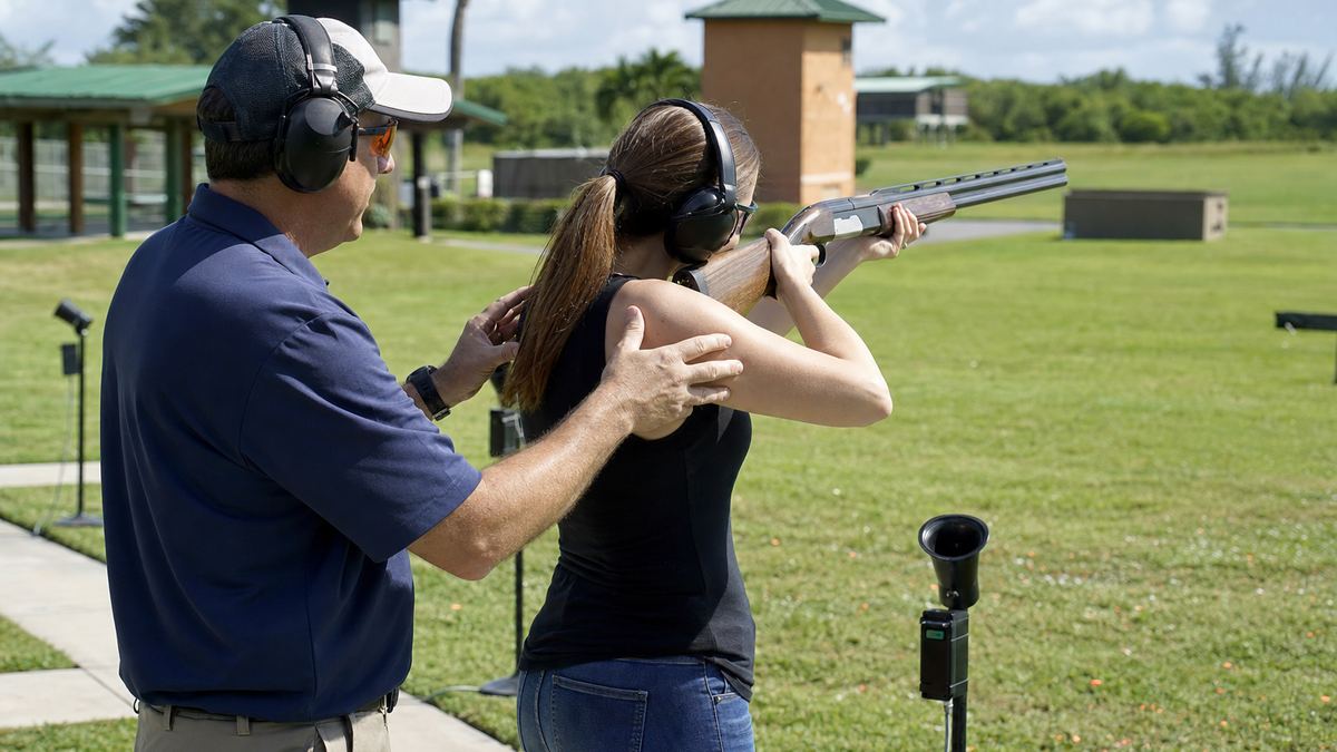 An instructor standing beside a beginner on a shotgun range, coaching her stance as she shoulders a shotgun, both wearing safety glasses and ear protection