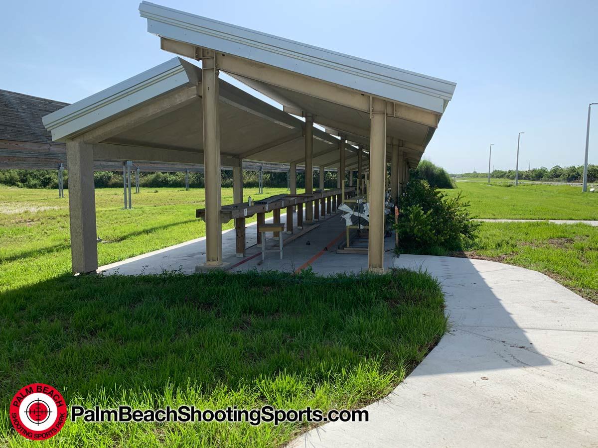 A long covered firing line with a peaked white roof and a concrete walkway leading to it across the grass