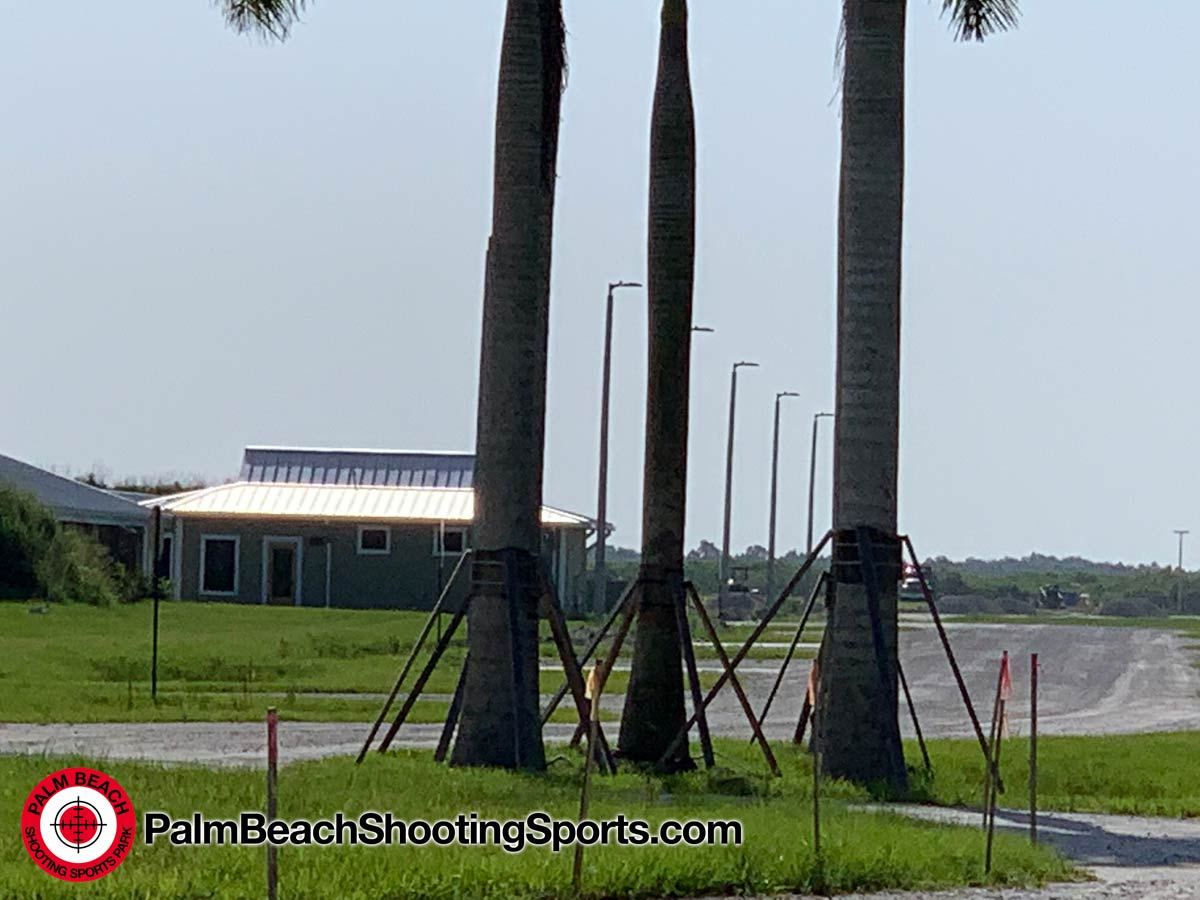 A pale green range building behind a row of newly planted palm trees, seen from the entrance gate