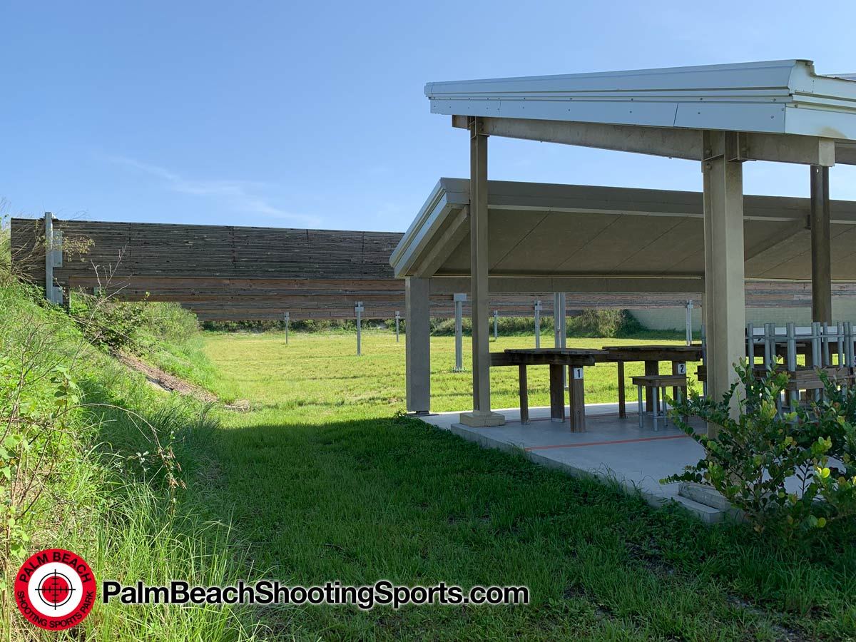 A covered firing line next to a grass range with target frame posts and a tall wooden baffle wall