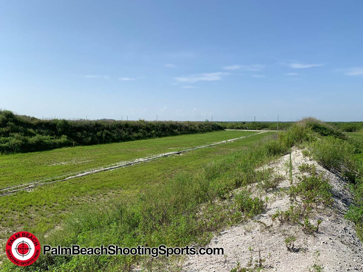 A wide grassy range seen from the top of an earth berm, with sandy soil in the foreground