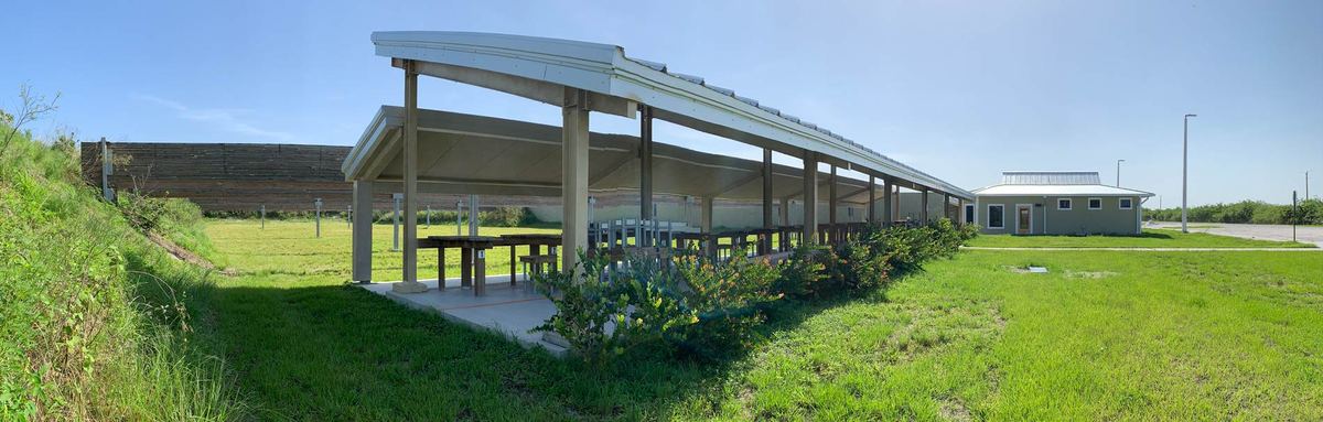 Panorama of a covered firing line with a curved metal roof beside a grassy earth berm, with the pale green range building at the far end